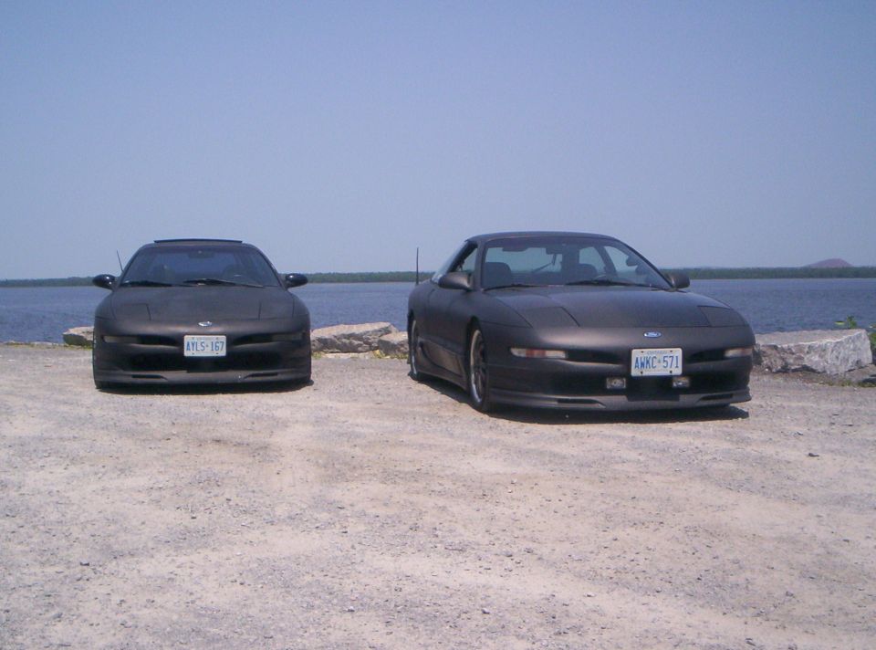 Two matte-black Ford Probe coupes parked on gravel by water