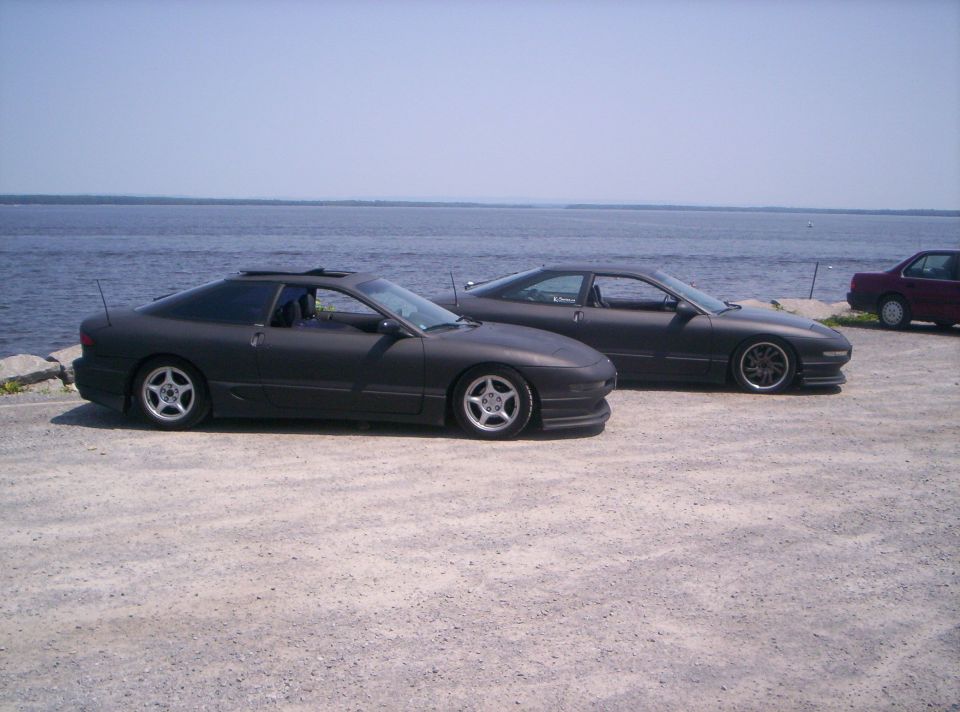 Two matte-black Ford Probe coupes parked on gravel by water