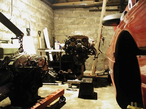 1992 Ford Taurus - Cinder-block garage mid-engine-swap: one V6 engine hangs from a hoist chain at left while a second V6 with intake and accessory belt drive…