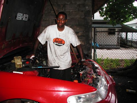 1992 Ford Taurus - A person in a dirt-smudged Geek Squad T-shirt stands in the engine bay opening of a red sedan with the hood raised, the underside of the ho…