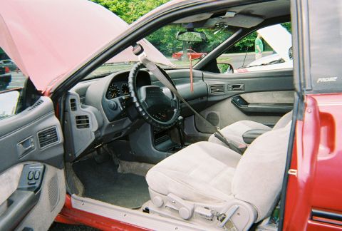 1991 Ford Probe - Interior of a red first-generation Ford Probe shot through the open passenger door at an outdoor show, showing gray cloth bucket seats, a m…