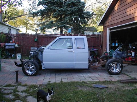 1998 Ford Taurus - A light blue pickup truck chassis sits on a paved driveway next to a garage, awaiting restoration.
