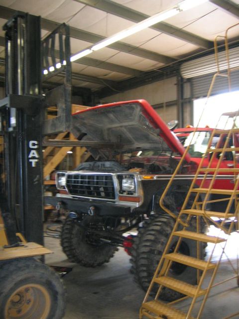 1979 Ford Bronco - A red 1979 Ford Bronco (or similar era Ford) with the hood/front clip tilted up and a lift kit sits inside a metal-frame workshop, with a y…