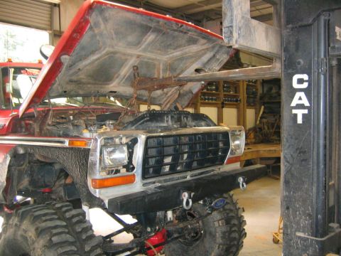 1979 Ford Bronco - A lifted red Ford 4x4 sits in a shop with its hood raised and the engine bay empty, a chain hoist hanging over the open engine compartment…