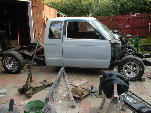 Pickup truck body on a chassis with engine parts visible. It's on jack stands outside a garage