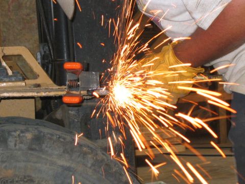 1979 Ford Bronco - A gloved worker in a white T-shirt grinds a steel bracket clamped in orange-handled C-clamps to a frame rail, throwing a heavy spray of ora…