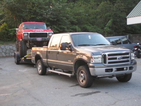 2005 Ford F-250 - A gray Ford F-250 tows a red Ford Bronco on a trailer, with a dark SUV and motorcycle visible in the background.