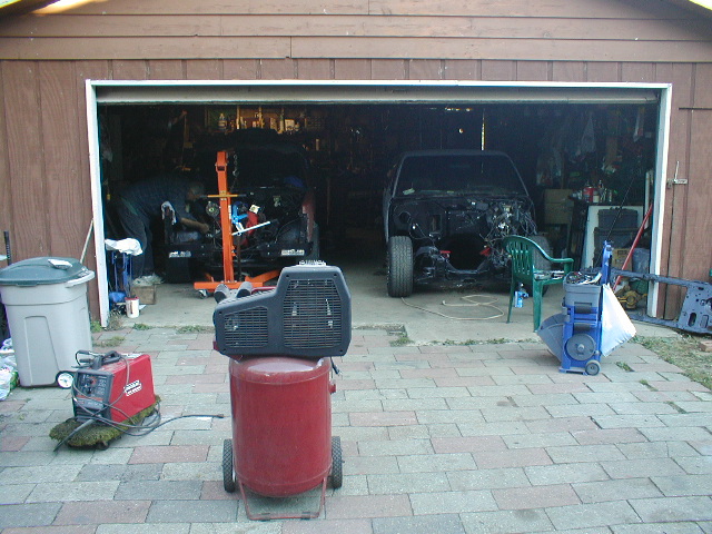 Two cars in a garage, one with an engine hoist, the other stripped down