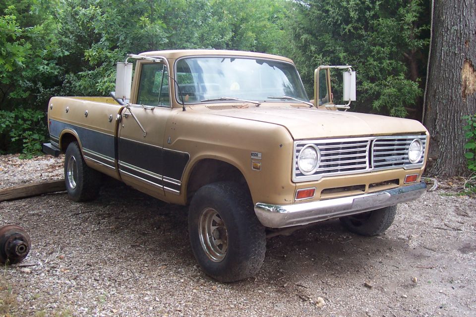 Tan vintage International pickup truck with dark lower trim and dual mirrors, parked on gravel by trees