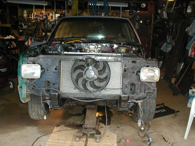 Vehicle front end disassembled with radiator and fan exposed, supported by jacks in a garage