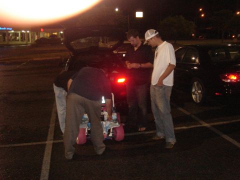 1996 Ford Probe - Several people are gathered around a cart with pink wheels, with a black car visible in the background at night.