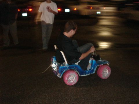 1996 Ford Probe - A person sits in a blue toy jeep with pink wheels on a wet road at night.