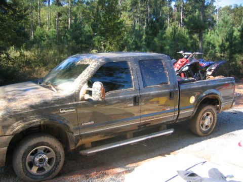 2005 Ford F-250 - Muddy 2005 Ford F-250 Super Duty pickup truck carries two ATVs in its bed, parked outdoors with trees behind.