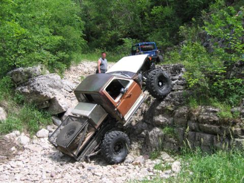 1981 Ford Bronco - Two off-road vehicles navigate a challenging rocky trail in a wooded area.
