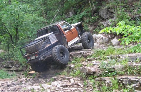 1981 Ford Bronco - A heavily modified off-road vehicle navigates a challenging rocky incline in a wooded area.