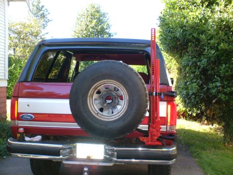 1989 Ford Bronco - Rear view of a red SUV, likely a 1989 Ford Bronco, equipped with a spare tire and a Hi-Lift jack mounted on the tailgate.