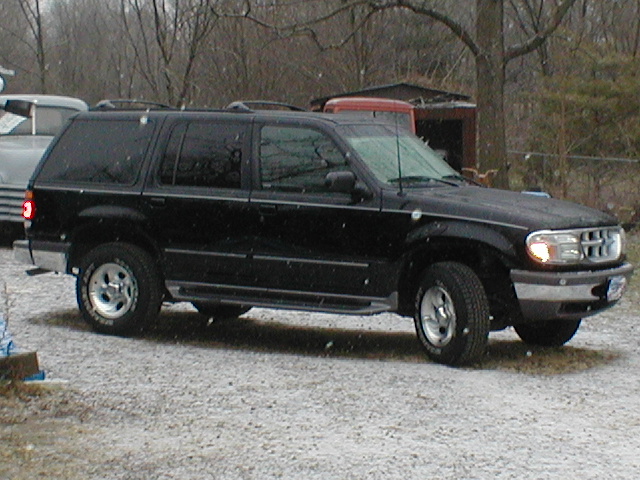 Black SUV parked outside with snow falling. Another vehicle is partially visible to the left