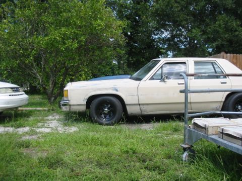 1985 Ford LTD Crown Victoria - A weathered sedan sits parked on a grassy lot, with another vehicle partially visible to its left.