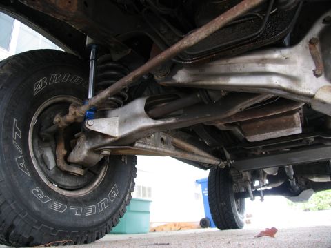 1984 Ford Bronco - Low underbody view of a lifted truck's Twin Traction Beam front suspension, showing the beam, radius arm, coil spring and a blue-bodied sho…
