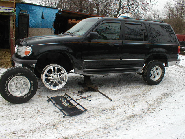 Black Ford Explorer on jack, front wheel removed, spare wheel and jack on ground