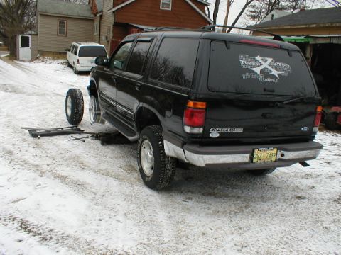 1998 Ford Taurus - A black Ford Explorer SUV is jacked up in the snow with one wheel removed, and a white van is parked nearby.