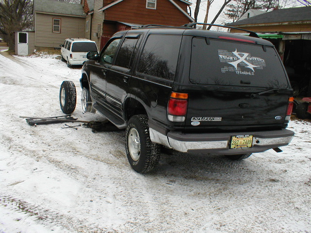 Black Ford Explorer SUV, rear view, jacked up with a wheel removed, snow on ground