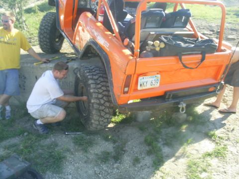 1994 Ford Bronco - A man works on the tire of an orange off-road vehicle with a roll cage and custom bumper.
