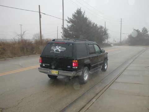 1998 Ford Taurus - A black SUV is seen from the rear on a wet road with trees and power lines in the background.