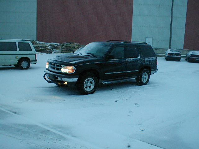 Black Ford Explorer parked in snow, front end visible. White van to the left, other cars in background