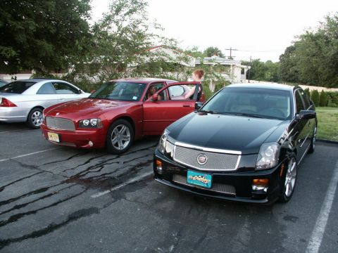 2001 Mercury Grand Marquis - Two sedans, a black Cadillac and a red Dodge, are parked side-by-side in a parking lot with trees and buildings in the background.
