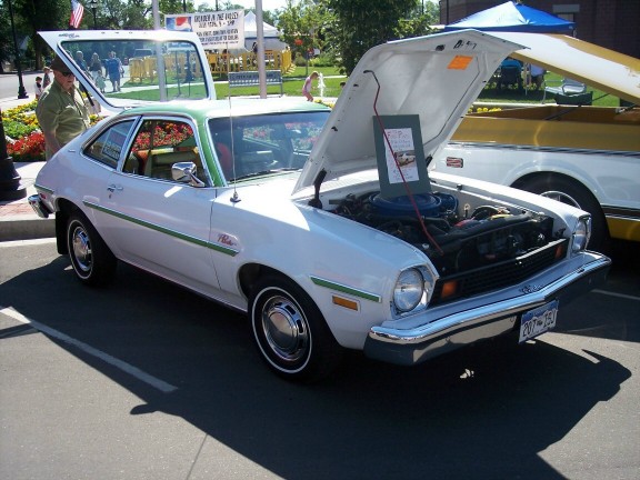 White 1970s Ford Pinto sedan with green trim, hood open, parked next to a yellow vehicle