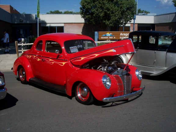 Red hot rod coupe with flames painted on the side, hood open showing engine. Another car is parked next to it
