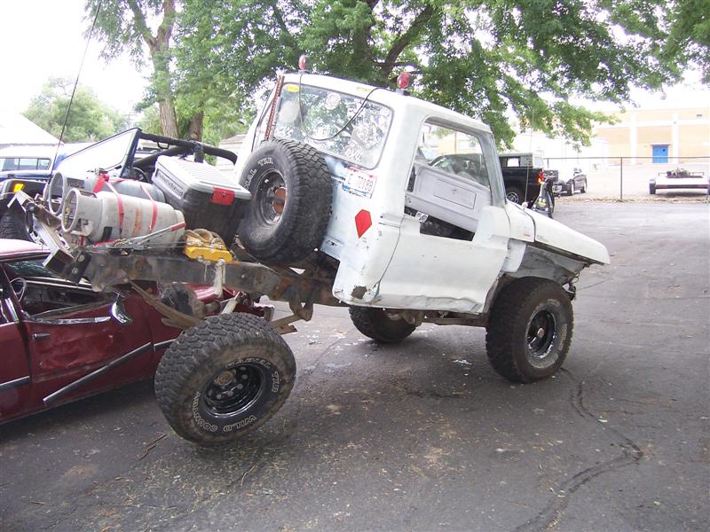 White pickup truck chassis with spare tire mounted on rear, propane tank and other items loaded. Another vehicle is partially visible underneath
