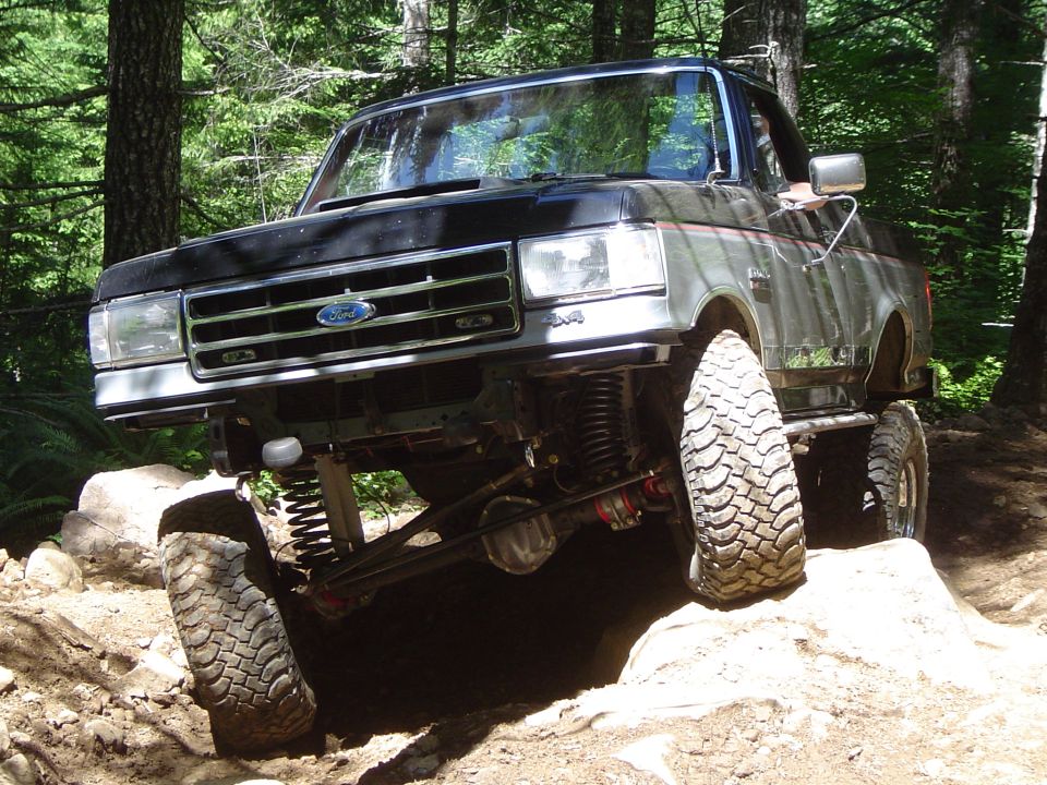 Lifted black Ford Bronco with exposed front axle flexing over rocks on a forest trail