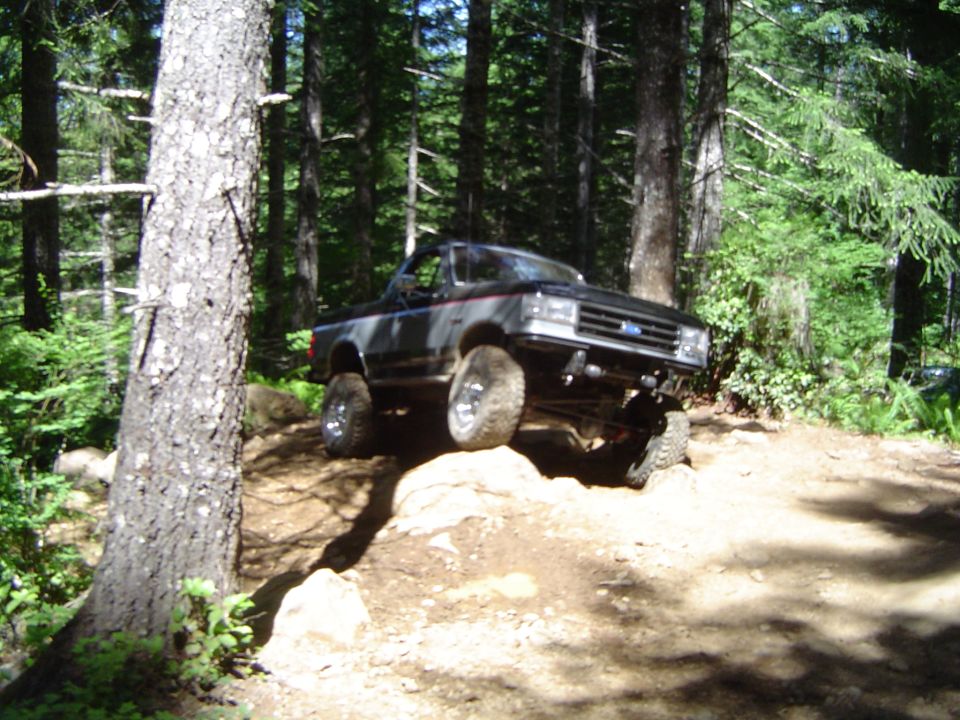Lifted black Ford Bronco flexing over a rock ledge on a forest trail