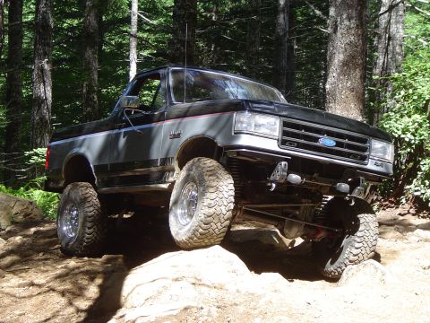 1989 Ford Bronco - A modified Ford Bronco navigates a rocky off-road path in a forest setting.