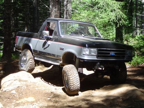 1989 Ford Bronco - A two-tone grey and black 1989 Ford Bronco navigates a challenging rocky trail in a forest setting.