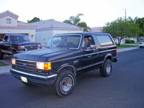 1991 Ford Bronco - A black Ford Bronco drives down a suburban street, with another Ford truck in the background.