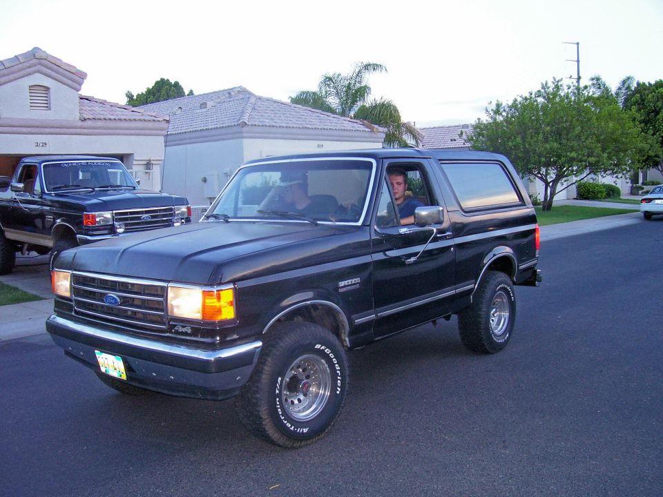 Black Ford Bronco driving on asphalt street, front 3/4 view. Driver visible. Another Ford truck parked in driveway