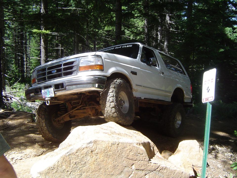 White Ford Bronco climbing over a large rock on a wooded trail. The vehicle has off-road tires and a lifted suspension