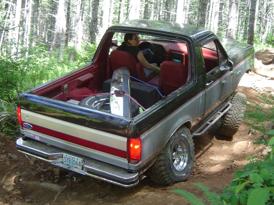 Black and silver Ford Bronco with top removed on a rutted forest trail, rear three-quarter view