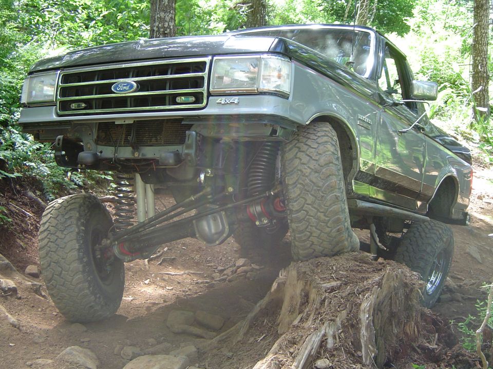 Lifted gray Ford 4x4 truck climbing tree stump on wooded trail, front axle flexed