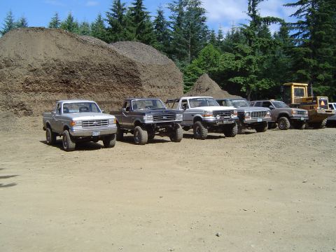 1989 Ford Bronco - Several lifted trucks and SUVs are parked in a dirt lot, with large mounds of dirt and trees in the background.