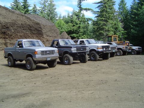 1989 Ford Bronco - A row of modified Ford trucks and SUVs are parked outdoors near a construction site.