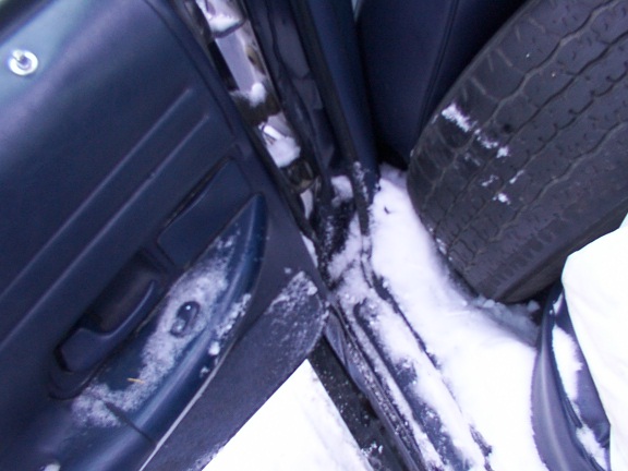 Open car door over snow showing damaged door edge and tire, low-angle ground view