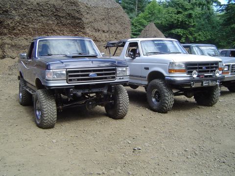 1989 Ford Bronco - Two modified Ford trucks, likely Broncos or F-Series, are parked side-by-side on a dirt lot.