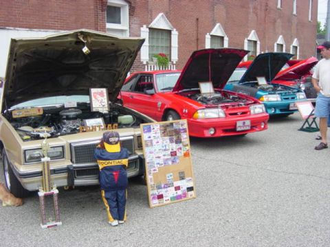 1995 Ford Probe - A car show features several vehicles with their hoods open, attracting attention from attendees and a young admirer.