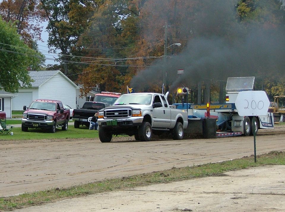 Silver Ford Super Duty truck pulling a sled, emitting black smoke. Another red Ford truck is parked nearby