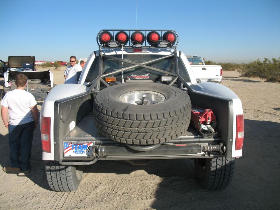Rear view of a white off-road truck with a spare tire mounted in the bed, roll cage, and lights on top. Another white truck is visible in the background