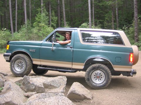 1990 Ford Bronco - A green and tan Ford Bronco navigates a rocky trail in a wooded area.